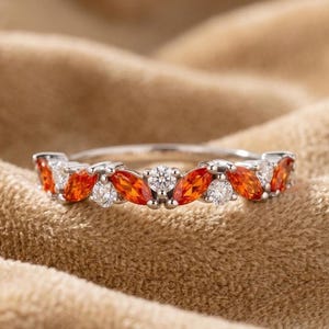 May include: A silver ring with alternating orange and white gemstones. The orange stones are marquise-cut, and the white stones are round. The ring is set against a tan background.