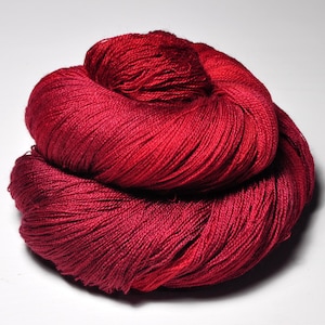 May include: A tightly wound skein of red yarn, with varying shades from deep burgundy to bright crimson. The yarn appears soft and finely spun, suitable for knitting or crochet projects. The background is a plain white surface.