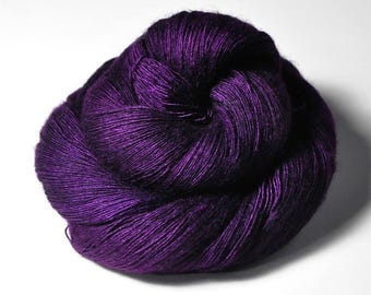 May include: A skein of deep purple yarn with a distinct sheen and a tightly twisted texture. The yarn is wound into a rounded, compact shape, showcasing a rich, dark violet colour with subtle highlights that emphasise the individual fibre strands.