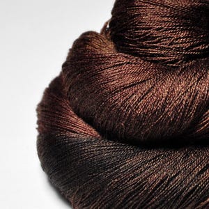 May include: A close-up of a skein of brown yarn with a gradient effect, transitioning from dark brown to a lighter shade. The yarn is tightly wound, showing the texture of the fibers. Ideal for knitting and crochet projects.