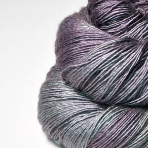 Puede incluir: Primer plano de un ovillo de hilo en tonos grises, morados y turquesas. El hilo está bien enrollado, con hebras visibles y un aspecto suave y texturizado. El degradado de color crea un efecto sutil y artístico, ideal para proyectos de punto o ganchillo.