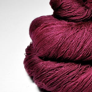 Pode incluir: Close-up de um novelo de fio bordô escuro. O fio é enrolado firmemente, mostrando uma superfície texturizada. A cor é um tom rico e saturado de vermelho-violeta, ideal para projetos de tricô ou crochê.
