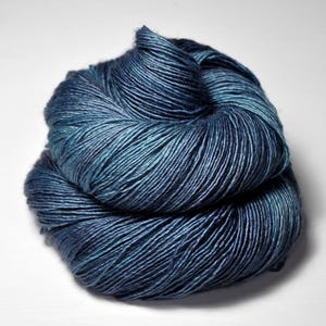 May include: A skein of yarn in shades of blue and teal. The yarn is tightly wound, with visible strands creating a textured appearance. The color transitions from dark blue to lighter teal hues.