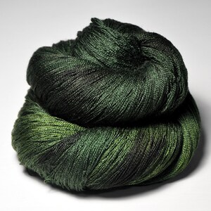 Peut inclure: Pelote de fil vert foncé, enroulée en boule. Le fil présente un léger éclat, avec des nuances de vert foncé et clair. La texture du fil est visible, suggérant une fibre naturelle et douce. Idéal pour les projets de tricot ou de crochet.