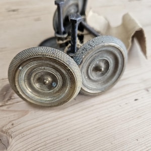 Op de afbeelding: Antieke metalen boekpers met twee cirkelvormige, getextureerde platen. De platen hebben een spiraalvormig ontwerp en zijn verbonden door een schroefdraadstang en een metalen frame. De pers is gemaakt van verouderd metaal en staat op een houten oppervlak.