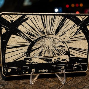 May include: A rectangular patch featuring a high-contrast black and beige graphic illustration of a cockpit view looking out into space with light streaks radiating from a central point, framed by a stitched border and supported by a clear stand.