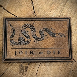 May include: A rectangular brown leather patch featuring a debossed graphic of a segmented snake and the text JOIN, or DIE. The snake is divided into eight parts, each labeled with letters representing colonial regions, set against a textured surface.