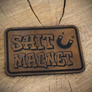 May include: A rectangular brown leather patch with rounded corners and a black border. The text SHIT MAGNET is embossed in a bold, blocky font, accompanied by a small black horseshoe magnet icon positioned to the right of the word SHIT.