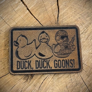 May include: A rectangular patch with a brown background and black border. The patch features two duck silhouettes and a third duck in military gear holding a gun. The text "DUCK, DUCK, GOONS!" is below the ducks.