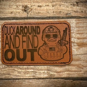 Può includere: Una toppa in pelle marrone con la scritta "DUCK AROUND AND FIND OUT" e l'illustrazione di un'anatra con equipaggiamento tattico e un fucile. La toppa è di forma rettangolare con angoli arrotondati.