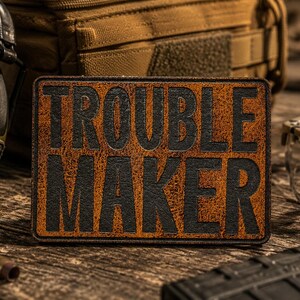 May include: A rectangular patch with a textured, distressed brown background and bold, black embossed lettering that reads TROUBLE MAKER. The patch features a clean, stitched border and a flat, durable surface design suitable for tactical gear.