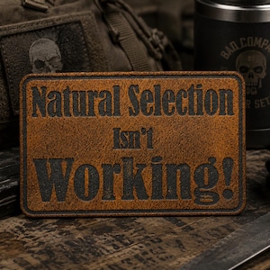 May include: A rectangular brown leather patch with a dark border and black embossed text that reads Natural Selection Isn't Working! The patch has a textured surface and rounded corners, featuring a bold, sans-serif font for the lettering.