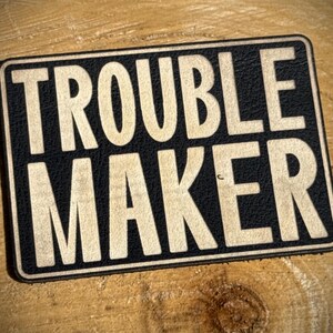 May include: A rectangular black and tan patch with the words "TROUBLE MAKER" in bold, white letters. The patch has a textured appearance and is set against a wooden background.
