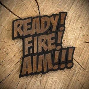 May include: A decorative sign featuring the text READY! FIRE! AIM!!! in a bold, block-style font. The letters have a textured, brown, wood-grain appearance and are outlined with a thick, solid black border, creating a layered, dimensional effect.