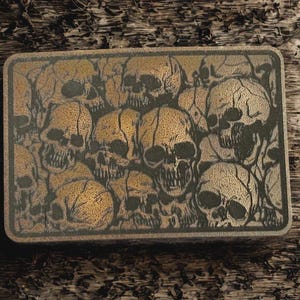 May include: Rectangular, bronze-colored metal box featuring a detailed pattern of skulls. The skulls are in varying sizes and orientations, creating a dense, textured design. The box has a dark border and a distressed, aged appearance.