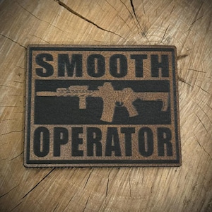 May include: A rectangular patch with the words "SMOOTH OPERATOR" in bold, black letters. A silhouette of a rifle is centered between the words. The patch has a distressed, brown leather-like appearance.