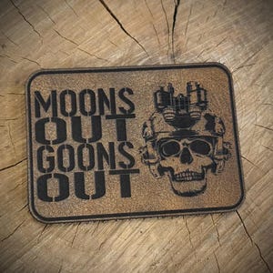 May include: A rectangular patch with the words "MOONS OUT GOONS OUT" in bold, black letters. A skull wearing a helmet with night vision goggles is on the right side. The patch has a textured, brown background.
