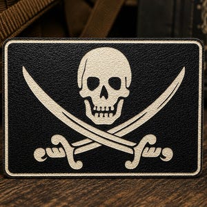 May include: A rectangular black patch featuring a cream-colored graphic of a human skull positioned above two crossed swords. The design is centered on the patch, which has a thin cream-colored border and a textured surface finish throughout.