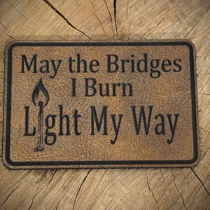 May include: A rectangular brown leather patch with a black border featuring the engraved text "May the Bridges I Burn Light My Way" and a graphic of a lit matchstick. The patch has a textured surface and rounded corners, displaying clear black lettering.