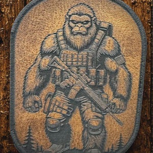 May include: A brown and black patch featuring a Sasquatch figure in tactical gear, holding a rifle. The design includes a detailed depiction of the creature's fur, gear, and the weapon. The patch has a textured appearance and a dark border.