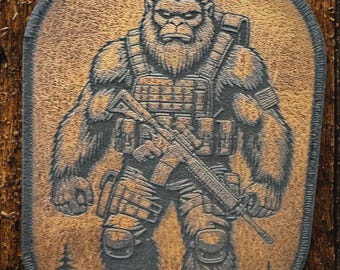May include: An oval-shaped patch featuring a dark, engraved illustration of a hairy, ape-like creature standing in a forest. The creature wears tactical gear, including a vest and knee pads, and holds a rifle across its chest with both hands.