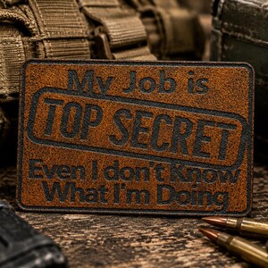 May include: A rectangular brown leather patch with black engraved text that reads: My Job is TOP SECRET Even I don't know What I'm Doing. The patch features a stitched border and a textured surface, presenting a clear and legible message in a bold font.