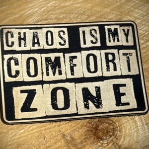 May include: A rectangular sign with the text "CHAOS IS MY COMFORT ZONE" in a distressed, vintage style. The words are in a bold, sans-serif font, with a black background and off-white lettering. The sign is set against a wooden background.