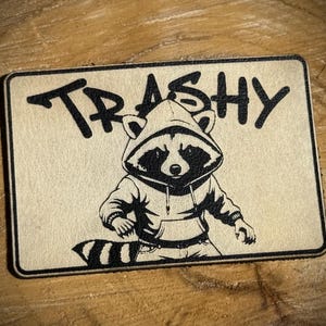 Könnte beinhalten: Rechteckiger Aufnäher mit einem Waschbären mit Kapuzenpullover und dem Wort "TRASHY" darüber. Der Waschbär ist schwarz-weiß, vor einem hellbraunen Hintergrund mit schwarzem Rand.