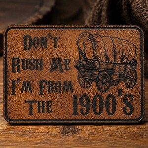 May include: A rectangular brown leather patch featuring a dark engraved illustration of a covered wagon and the text "DON'T RUSH ME I'M FROM THE 1900'S" in a bold, vintage-style font. The patch has a textured surface and a dark, defined border.
