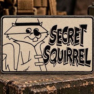 May include: A rectangular patch with a tan background and a black border. The design features a black line art illustration of a squirrel wearing a fedora hat and trench coat, accompanied by the black text "SECRET SQUIRREL" in a bold font.