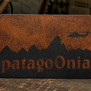 May include: A rectangular leather patch featuring a textured brown upper section and a black lower section. The design depicts a mountain range silhouette with a small helicopter icon above it. The text patagoOnia is engraved in the black area.