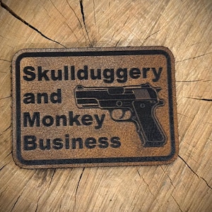 May include: A rectangular, brown leather patch with the words "Skullduggery and Monkey Business" and a black handgun graphic. The patch is set against a textured, wooden background.