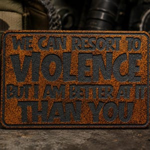 May include: A rectangular patch with a textured, distressed brown surface and black embossed lettering that reads: WE CAN RESORT TO VIOLENCE BUT I AM BETTER AT IT THAN YOU. The patch features rounded corners and a distinct, rugged appearance.