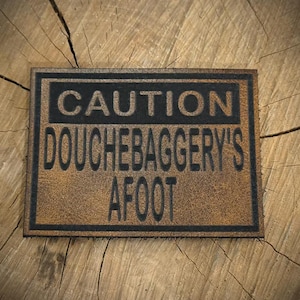 May include: A rectangular, brown leather sign with the words "CAUTION DOUCHEBAGGERY'S AFOOT" in black, bold letters. The sign has a distressed, vintage look and is set against a wooden background.