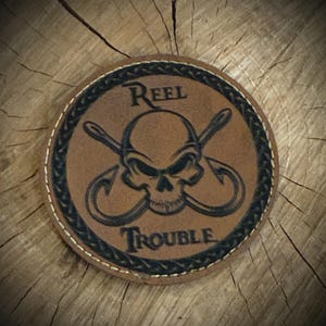 May include: Round, brown leather patch with a skull and crossed fishing hooks design. The words "Reel Trouble" are arched above and below the skull. The patch has a black braided border and white stitching. The patch is on a wooden surface.