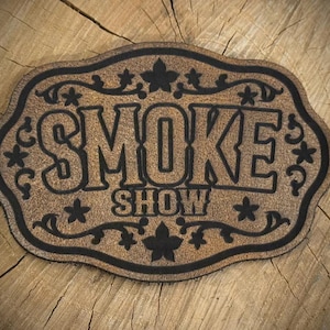 May include: A tan leather patch with a scalloped edge featuring the black embossed text SMOKE SHOW in the center. The design includes black decorative floral and vine motifs surrounding the lettering, creating a detailed and textured appearance.