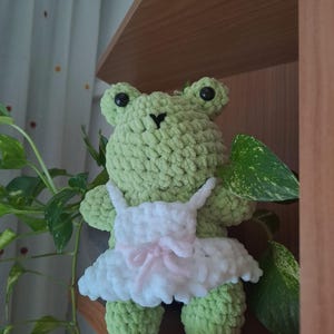 Può includere: Una bambola a forma di rana all'uncinetto verde chiaro fatta a mano con un vestito bianco con un fiocco rosa. La rana ha occhi neri a bottone ed è posizionata su una mensola di legno, con piante verdi e frondose sullo sfondo.