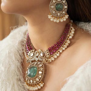 Puede incluir: Un conjunto de collar y pendientes ornamentados con grandes piedras de color esmeralda, rodeadas de diamantes y perlas más pequeños. El collar tiene una banda de color rubí y un colgante grande. Los pendientes reflejan el diseño del colgante.
