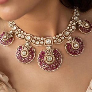 Op de afbeelding: Een goudkleurige ketting- en oorbellenset met een bloemmotief, met robijnkleurige stenen en heldere kristallen. De ketting heeft een centrale hanger en twee zijhangers, allemaal cirkelvormig. De oorbellen spiegelen het ontwerp.