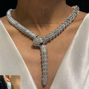 Puede incluir: Un collar de serpiente con incrustaciones de diamantes y pendientes a juego. El collar se envuelve alrededor del cuello y presenta una cabeza de serpiente con ojos de piedras preciosas verdes. Los pendientes reflejan el diseño del collar. La joyería está sobre un fondo blanco.