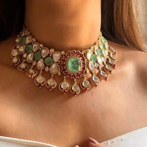 May include: A gold-toned choker necklace featuring emerald and ruby gemstones. The necklace has a central square emerald, surrounded by smaller rubies and teardrop-shaped diamond accents. Green and white stones are set in a floral pattern.