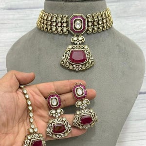 May include: A gold-toned jewelry set featuring a choker necklace, earrings, and a headpiece. The set includes ruby-colored gemstones and clear crystals. The necklace and earrings have a matching design, with a large ruby-colored stone in the center.