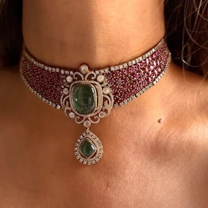 Pode incluir: Um luxuoso colar gargantilha com uma faixa larga de pequenas pedras preciosas vermelho escuro. O ponto focal é um grande pingente de pedra verde de corte quadrado, acentuado com pedras brancas menores e uma gota de pedra verde em forma de lágrima. O colar é cravejado em metal dourado.