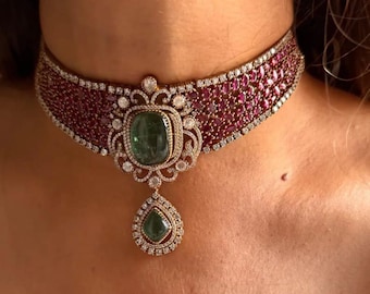 May include: A luxurious choker necklace featuring a wide band of small, deep red gemstones. The focal point is a large, square-cut green gemstone pendant, accented with smaller white gemstones and a teardrop-shaped green gemstone drop. The necklace is set in a gold-toned metal.