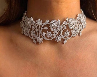 Op de afbeelding: Een zilverkleurige choker ketting met een ingewikkeld bloemenontwerp. De ketting heeft een reeks onderling verbonden bloem- en bladvormige elementen, bezet met sprankelende, heldere edelstenen. Het ontwerp is symmetrisch en wikkelt zich om de nek.