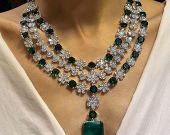 May include: A three-strand necklace featuring emerald-cut green gemstones and sparkling white diamonds. The necklace has a pendant with a large emerald-cut green gemstone. The necklace is worn on a person wearing a white top.