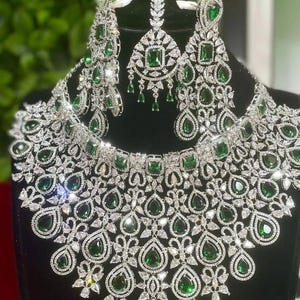 May include: A dazzling jewelry set featuring a necklace, earrings, and a headpiece. The set is silver-toned and adorned with sparkling clear and emerald-green gemstones. The necklace and earrings have a drop design, and the headpiece has a central pendant.