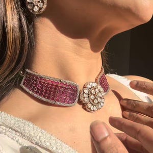 May include: A glamorous jewellery set comprising a choker necklace and earrings. The necklace has a rectangular, ruby-coloured centre with a diamond border. The earrings and necklace feature a floral design with clear and ruby-coloured stones.