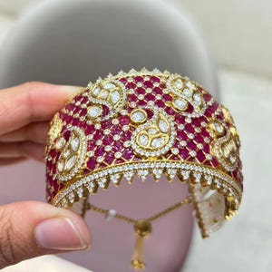 May include: A wide gold cuff bracelet featuring a dense lattice pattern set with numerous small red stones and clear white stones. The design includes several prominent paisley-shaped motifs outlined in white stones, with a delicate adjustable drawstring.