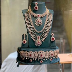 Puede incluir: Un conjunto de joyas completo exhibido en un soporte de terciopelo verde azulado. El conjunto incluye un collar, aretes y una pulsera, todos con diseños intrincados con piedras claras y acentos rojos.